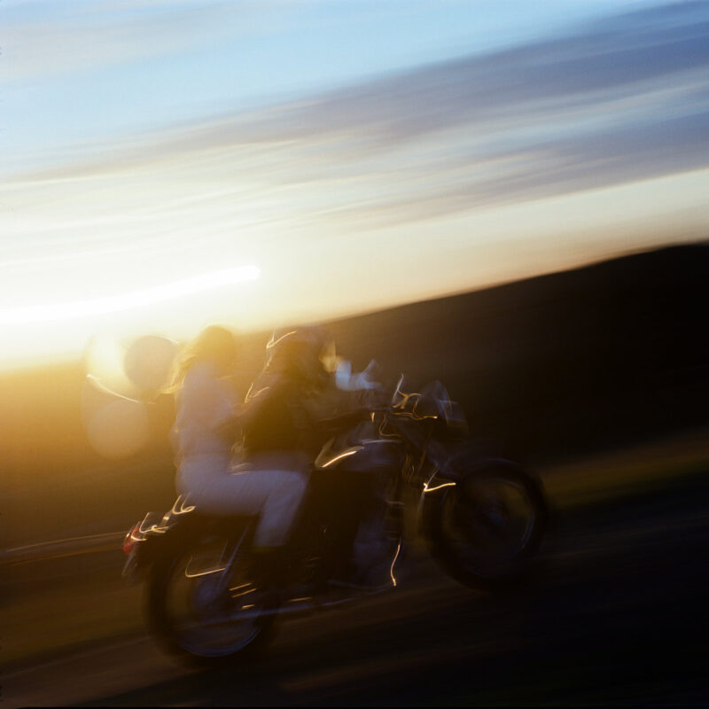 Verschwommene Aufnahme zweier Personen auf einem Motorrad bei Sonnenuntergang – ein Bild voller Tempo, Freiheit und emotionaler Aufladung.