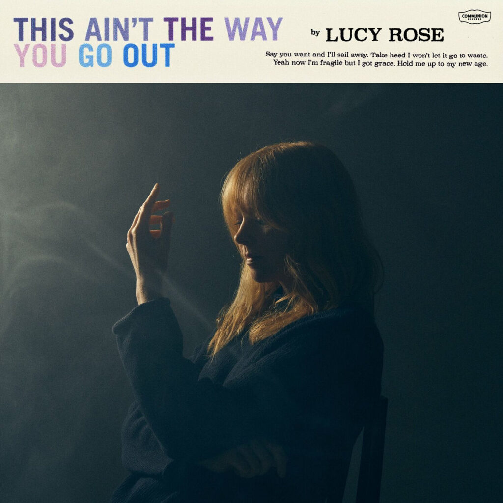 Lucy Rose in einem dunklen, rauchigen Raum, nachdenklich im Profil mit erhobener Hand – Cover des Albums „This Ain’t The Way You Go Out“.