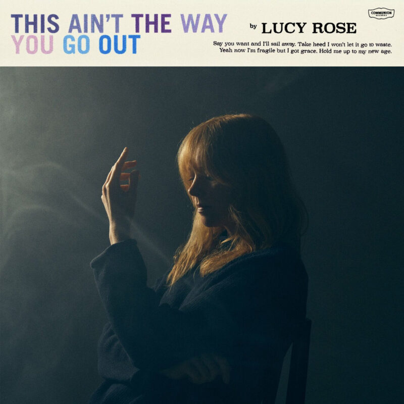 Lucy Rose in einem dunklen, rauchigen Raum, nachdenklich im Profil mit erhobener Hand – Cover des Albums „This Ain’t The Way You Go Out“.