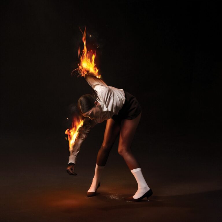 Eine Frau in Shorts, weißem Hemd und Pumps steht gebückt mit brennenden Armen – ein dramatisch stilisiertes Albumcover voller Symbolkraft.