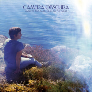 Eine Frau sitzt mit einem Buch auf einem Felsen am Wasser, umgeben von Heidekraut – Cover des Albums „Look to the East, Look to the West“ von Camera Obscura.