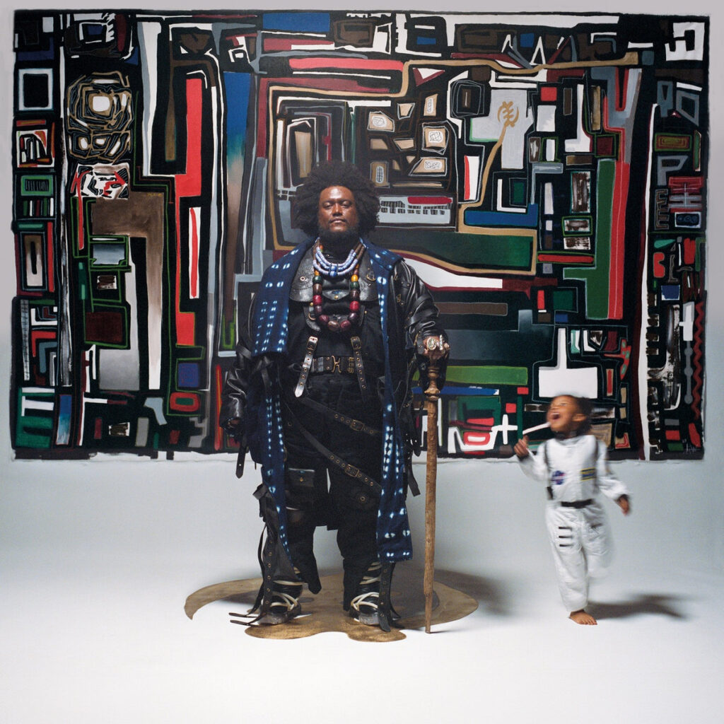 Cover des Albums „Fearless Movement“ von Kamasi Washington: Der Musiker steht in opulenter Kleidung vor einem farbenreichen abstrakten Wandgemälde, neben ihm ein lachendes Kind im Raumanzug.