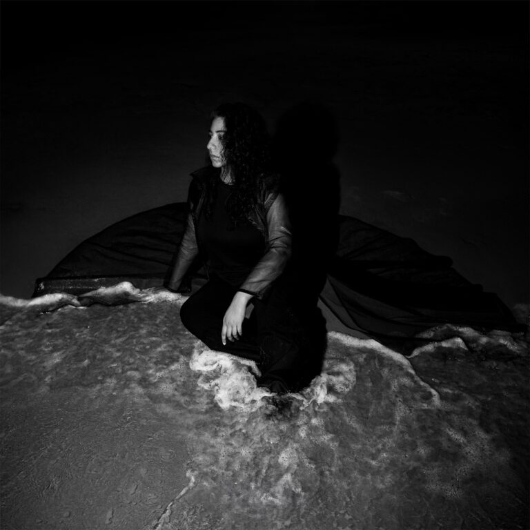 Schwarzweißcover von „Night Reign“: Arooj Aftab sitzt im Dunkeln am Ufer, ein langer Mantel fließt wie Schatten ins Wasser – still, stark, symbolisch.