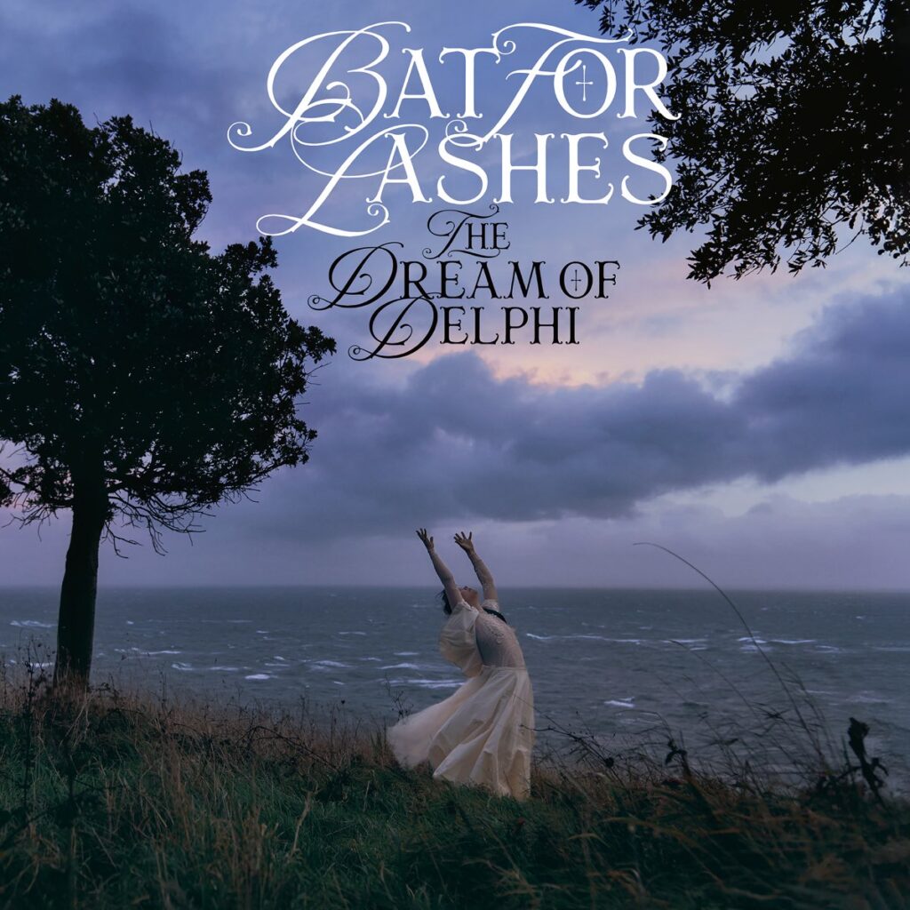 Eine in Weiß gekleidete Frau tanzt mit erhobenen Armen auf einer windgepeitschten Klippe am Meer, umgeben von dunklen Wolken – Cover zu Bat For Lashes’ The Dream of Delphi.