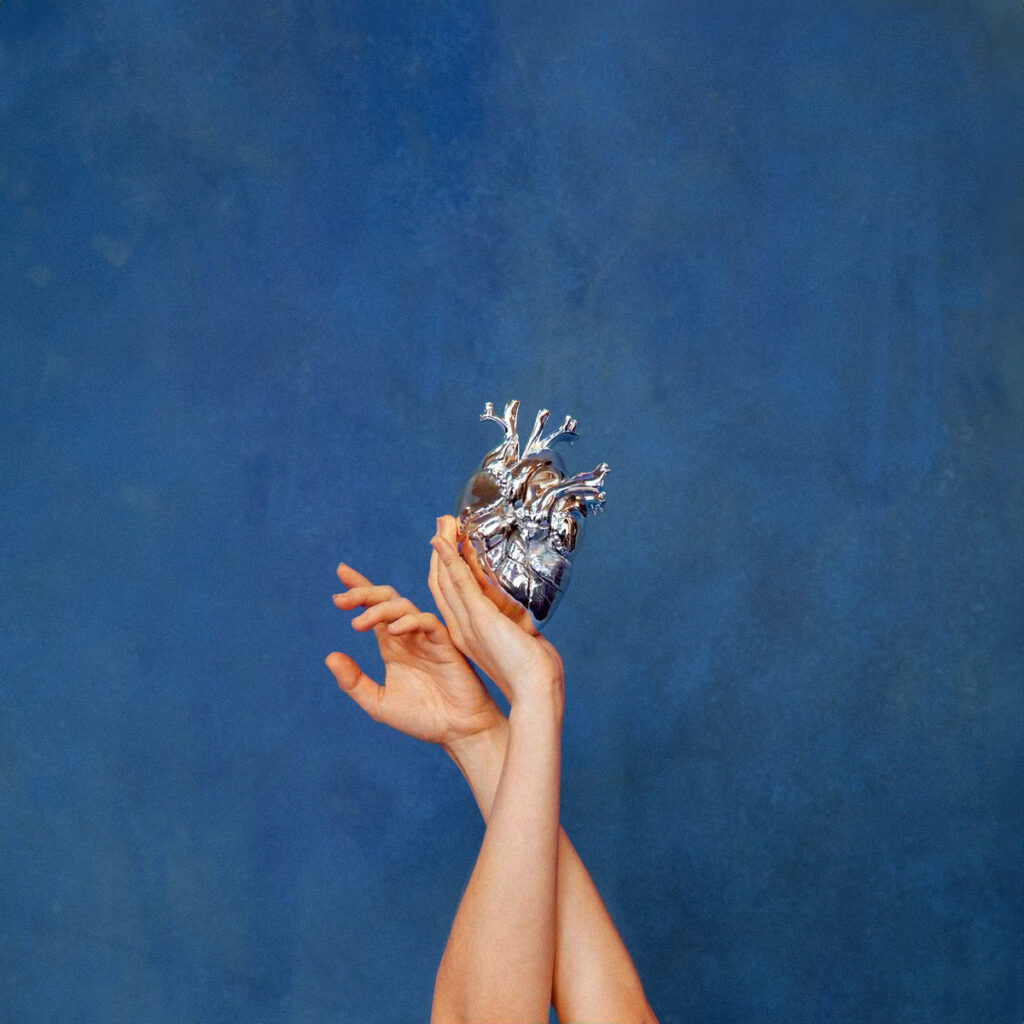 Zwei erhobene Hände halten ein silbernes, anatomisches Herz vor monochrom blauem Hintergrund – minimalistisches, symbolträchtiges Cover von AURORAs What Happened to the Heart?