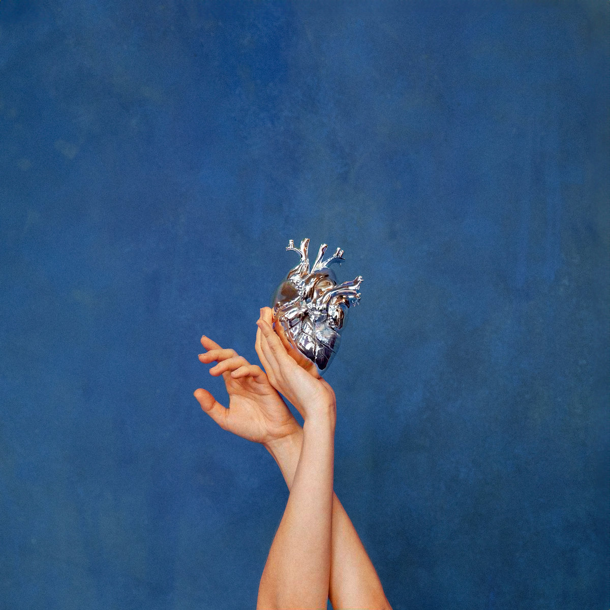 Zwei erhobene Hände halten ein silbernes, anatomisches Herz vor monochrom blauem Hintergrund – minimalistisches, symbolträchtiges Cover von AURORAs What Happened to the Heart?