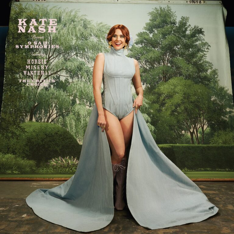 Kate Nash posiert lächelnd in hellblauem Kleid vor gemalter Gartenkulisse – das verspielte Cover des Albums „9 Sad Symphonies“.