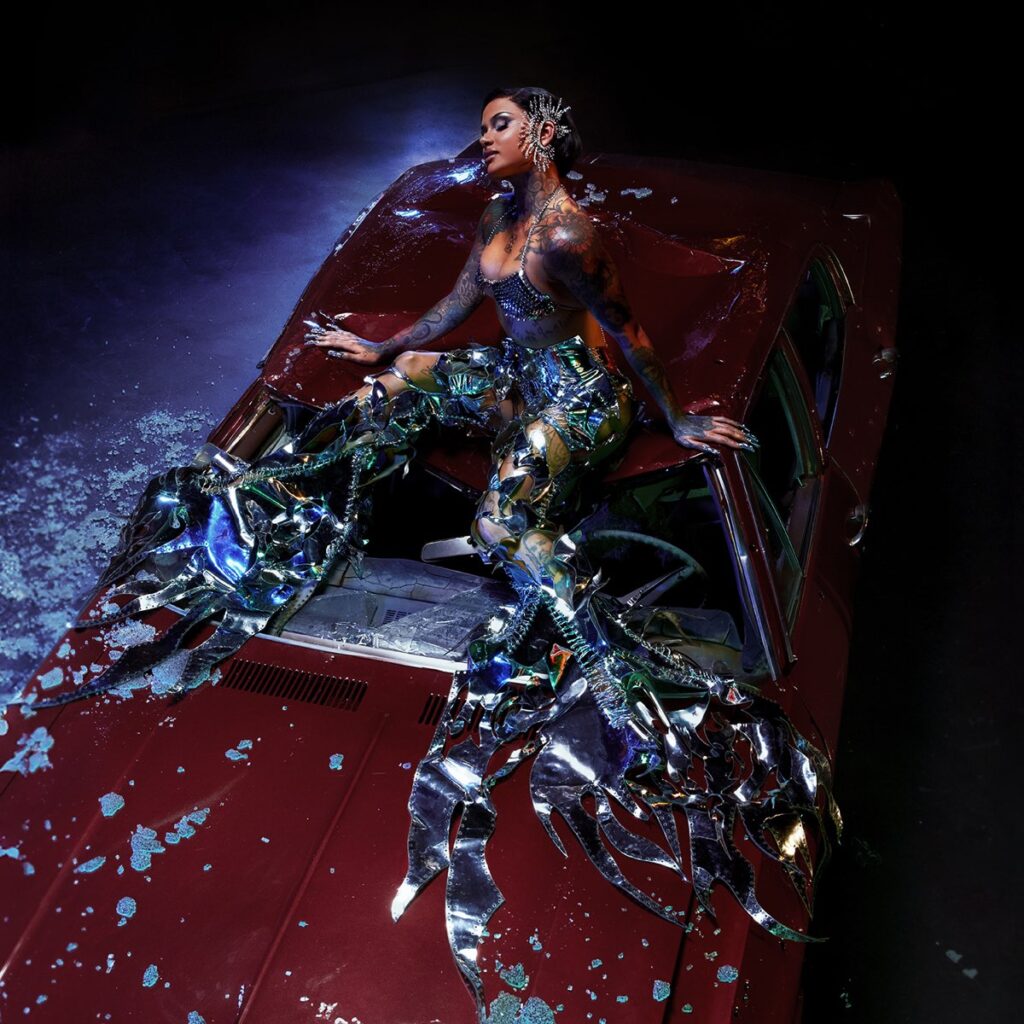 Kehlani sitzt in futuristischem, spiegelndem Outfit auf einem zerdellten, roten Auto – umgeben von Glassplittern und dramatischem Licht – visuell kraftvolles Cover ihres Albums CRASH.