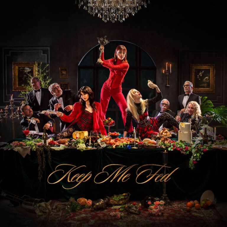 Das Cover von Keep Me Fed zeigt The Warning an einer üppig gedeckten Tafel, umgeben von älteren Herren in Anzügen – die Szenerie erinnert an ein dekadentes Barockbankett, das von rebellischer Frauenpower aufgebrochen wird.