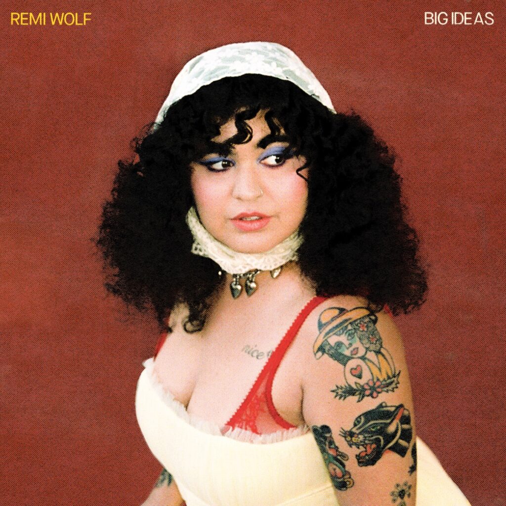Das Cover von Big Ideas zeigt Remi Wolf vor rotem Hintergrund in einem Vintage-Look mit Spitzenhaube, Tattoos und direktem Blick zur Seite – verspielt, stark, retro-modern.