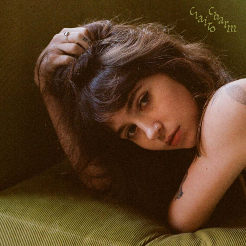 Clairo liegt mit verträumtem Blick auf einem olivgrünen Sofa, den Kopf in die Hand gestützt, mit sanftem Licht und dem Schriftzug „Charm“ in handschriftlicher Typo – minimalistisches Albumcover mit Vintage-Feeling.