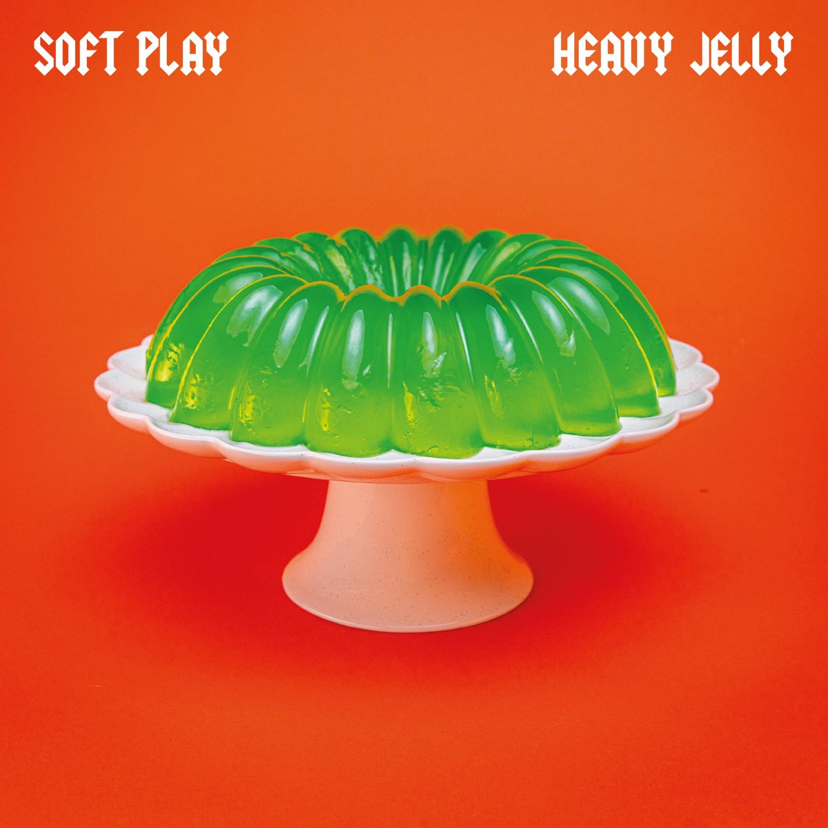 Grüne Wackelpudding-Kranzform auf weißer Tortenplatte vor knallorangefarbenem Hintergrund mit Schriftzug „Soft Play – Heavy Jelly“.
