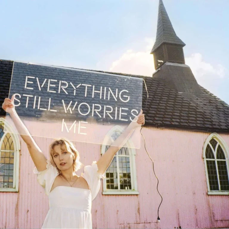 Abbie Ozard in weißem Kleid vor einer rosa Kirche, hält ein transparentes Schild mit der Aufschrift „Everything Still Worries Me“ – Albumcover ihres Debüts.