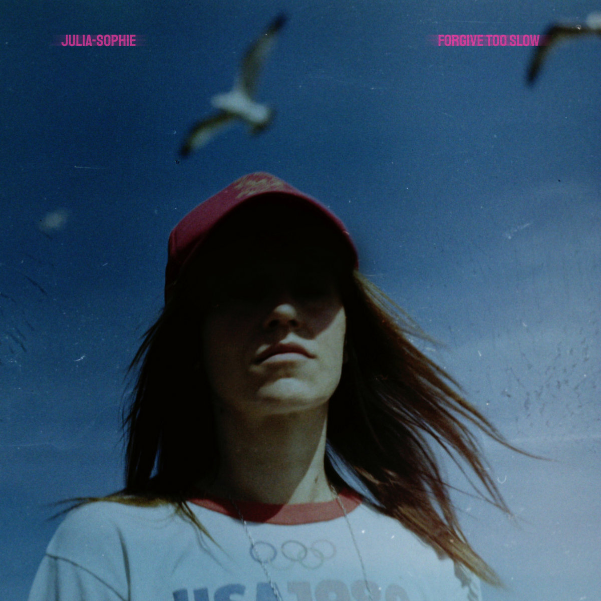 Das Cover von Forgive Too Slow zeigt Julia-Sophie unter einem blauen Himmel mit Möwen, ihr Gesicht im Schatten einer roten Kappe – ein stilles, introspektives Porträt zwischen Bewegung und Melancholie.