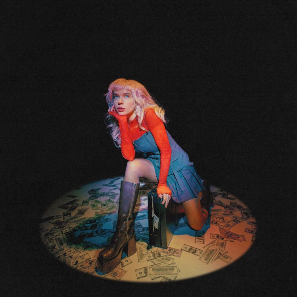 Pixey kniet in einem Lichtkegel auf einem Teppich aus Geldscheinen, trägt ein blaues Kleid und hohe Stiefel, umgeben von Dunkelheit und Drama – ein Bild zwischen Pop-Glamour und Kapitalismuskritik.