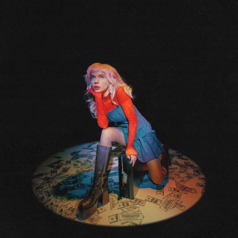 Pixey kniet in einem Lichtkegel auf einem Teppich aus Geldscheinen, trägt ein blaues Kleid und hohe Stiefel, umgeben von Dunkelheit und Drama – ein Bild zwischen Pop-Glamour und Kapitalismuskritik.