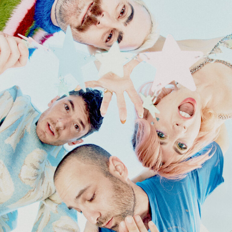 Buntes Bandfoto von Charly Bliss: Vier Bandmitglieder greifen lachend in die Kamera, mit Sternenketten vor hellblauem Hintergrund.