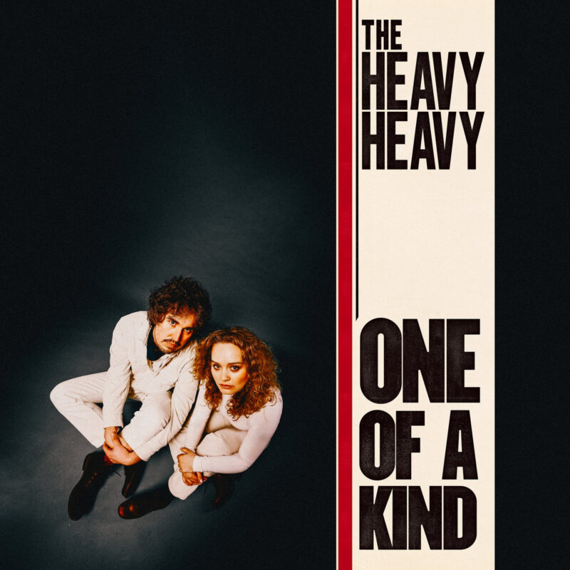 Albumcover zu "One Of A Kind" von The Heavy Heavy: Retro-inspiriertes Artwork mit dem Duo in weißem Outfit auf dunklem Boden, grafisch eingerahmt.