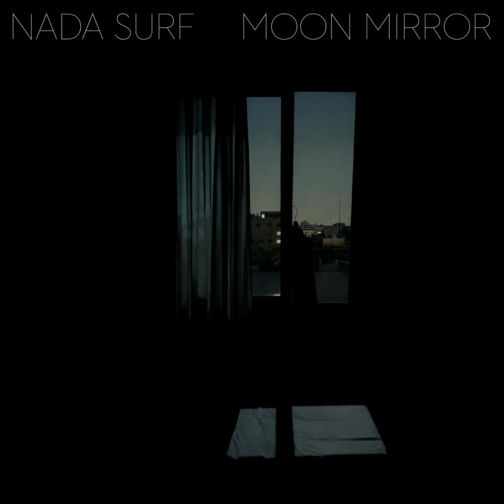 Albumcover zu "Moon Mirror" von Nada Surf: dunkles Zimmer mit Fensterblick bei Dämmerung, Stadtlichter im Hintergrund, melancholisch und ruhig.