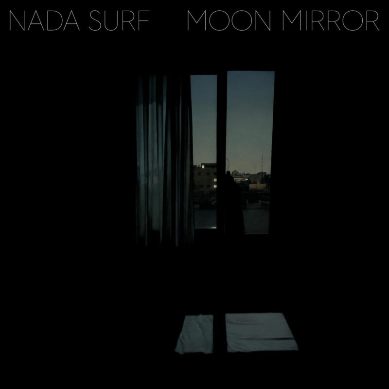 Albumcover zu "Moon Mirror" von Nada Surf: dunkles Zimmer mit Fensterblick bei Dämmerung, Stadtlichter im Hintergrund, melancholisch und ruhig.