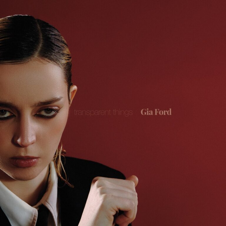 Nahaufnahme von Gia Ford in dunklem Anzug vor bordeauxfarbenem Hintergrund – Albumcover zu Transparent Things, mit entschlossenem Blick und nassem Haar.
