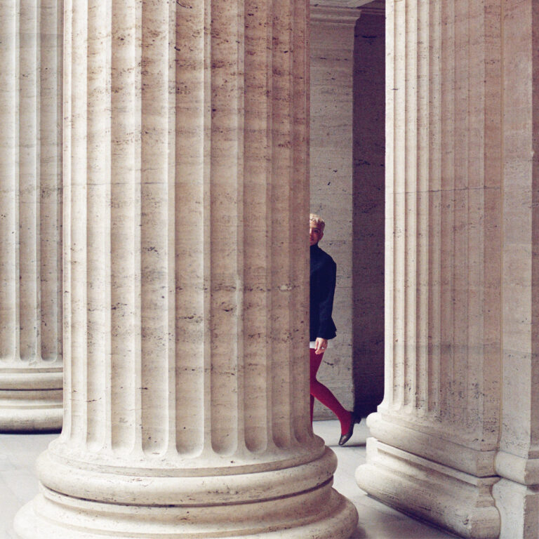 Cover von Tasha’s Album „All This and So Much More“: Eine Person in roter Strumpfhose tritt zwischen monumentalen Säulen hervor, teils verborgen, teils sichtbar.