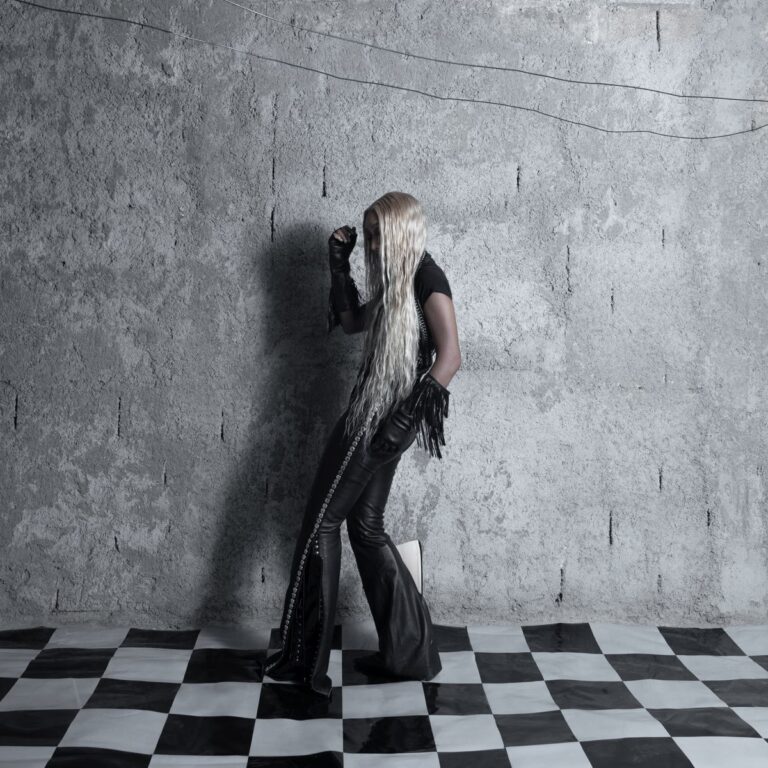 Cover von Fousheé’s Album „Pointy Heights“: Die Künstlerin mit langen blonden Haaren steht in schwarzem Leder-Outfit vor Betonwand auf Schachbrettboden – geheimnisvoll und stilisiert.