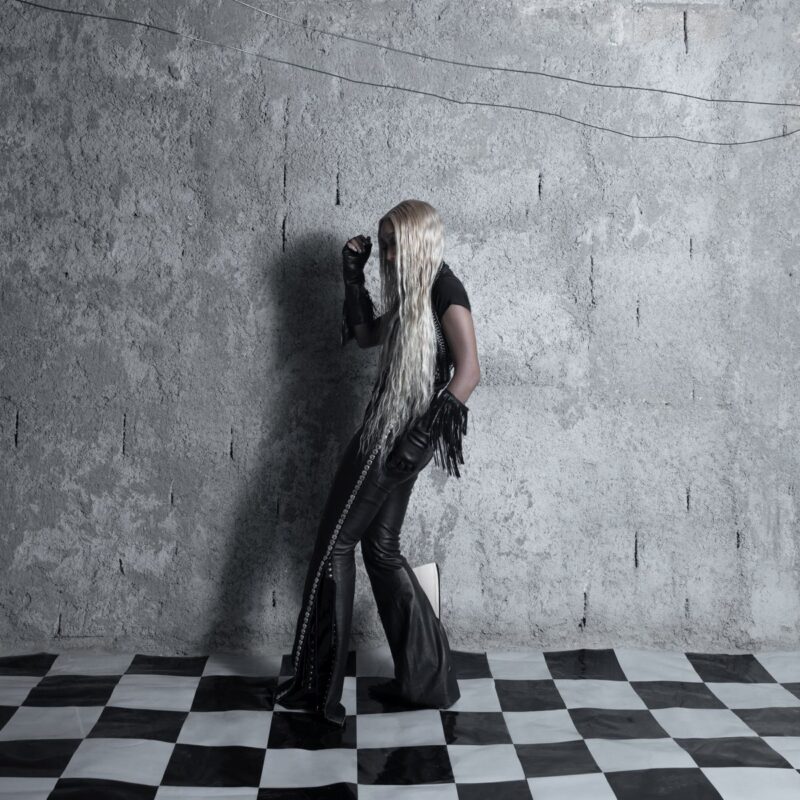 Cover von Fousheé’s Album „Pointy Heights“: Die Künstlerin mit langen blonden Haaren steht in schwarzem Leder-Outfit vor Betonwand auf Schachbrettboden – geheimnisvoll und stilisiert.