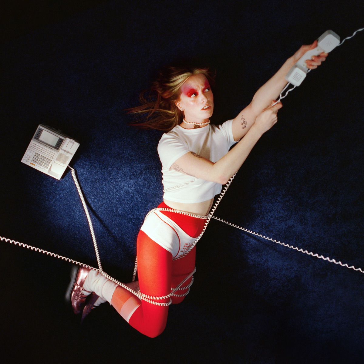 Orla Gartland liegt verkabelt mit Telefonhörern auf einem tiefblauen Teppichboden, in roter Strumpfhose und weißem Shirt – surreal, verspielt, ausdrucksstark.