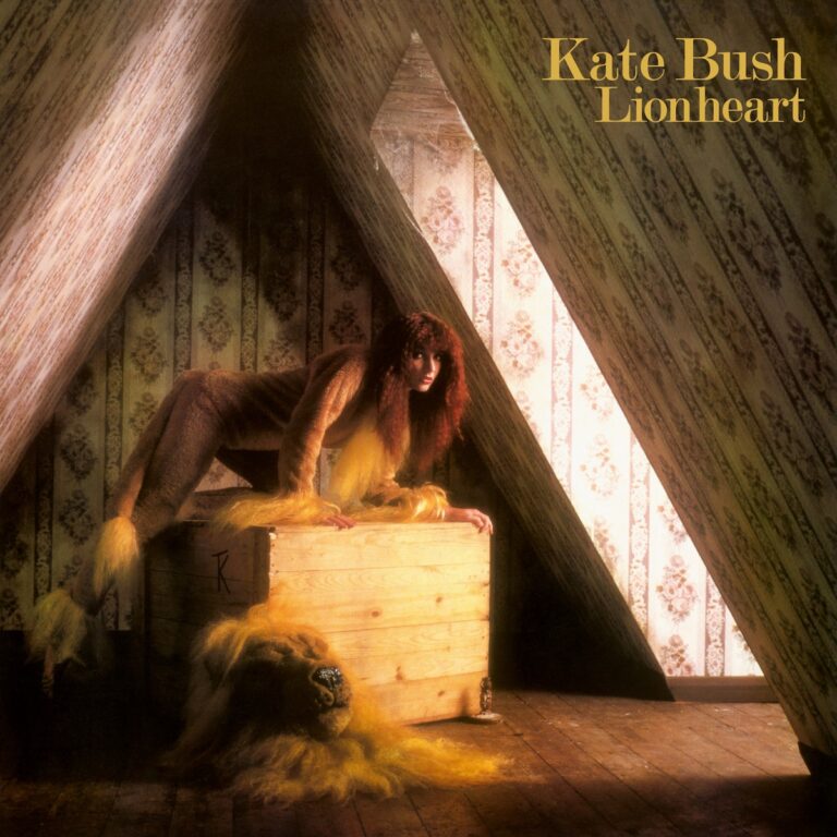 Kate Bush in einem braunen Kostüm mit Löwenmähne, auf einer Holzkiste in einem nostalgischen Raum.
