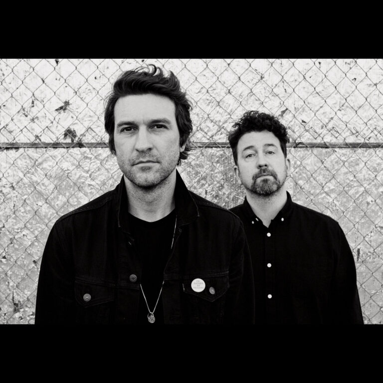 Schwarz-weiß-Foto von Japandroids vor Maschendrahtzaun: Zwei Männer mit ernster Miene, Symbol für die Reifephase der Band im Album Fate & Alcohol.