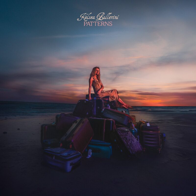 Kelsea Ballerini sitzt bei Sonnenuntergang auf einem Haufen Koffer am Strand – Cover des Albums Patterns, das emotionale Reise und Neubeginn symbolisiert.