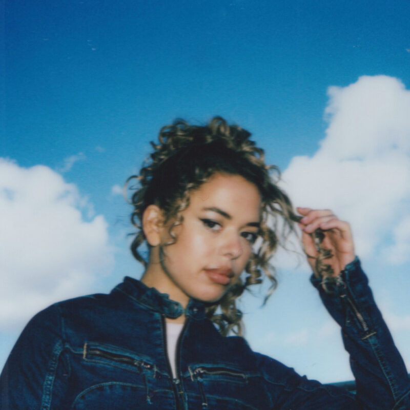 Porträt von Nilüfer Yanya vor strahlend blauem Himmel mit lockigem Haar und Jeansjacke – unscharf wie eine Polaroid-Aufnahme, verträumt und nostalgisch.