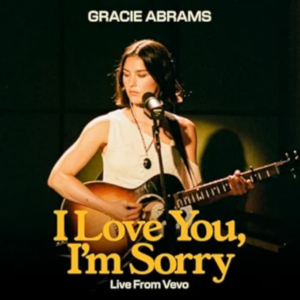Gracie Abrams sitzt mit Gitarre vor einem Mikrofon in stimmungsvollem Studiolicht, konzentriert und verletzlich – Szene aus ihrer Live-Session „I Love You, I’m Sorry“.