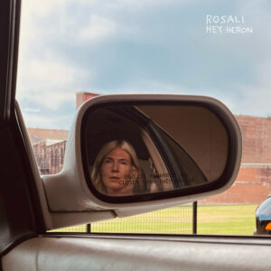 Cover des Songs „Hey Heron“ von Rosali: Blick aus dem Auto auf eine Frau im Rückspiegel, mit ruhiger Landschaft und Backsteingebäude im Hintergrund.
