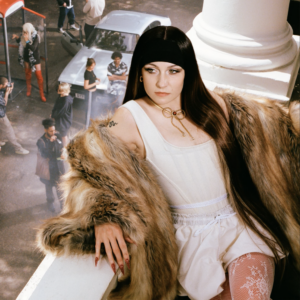 Cover zu Biig Piigs „Favourite Girl“: Die Künstlerin in weißem Outfit und Fellmantel auf einem Balkon, umgeben von Straßenszene – zwischen Luxus, Distanz und Pop-Drama.