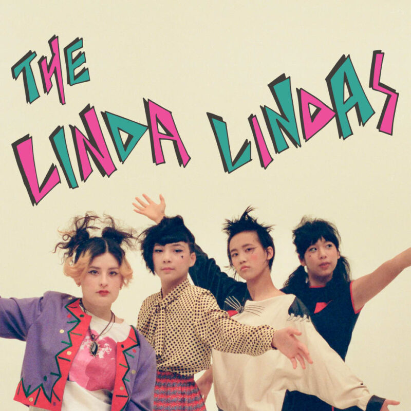 Die Band The Linda Lindas posiert energiegeladen vor hellem Hintergrund mit bunter Punk-Ästhetik und dem Schriftzug ihres Bandnamens.