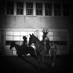 Samara Cyn sitzt selbstbewusst auf einem Pferd vor einer Garagenfassade bei Nacht, stilisiert im Western-Look mit Pelzmütze und Lederoutfit – Szene aus dem „Sinner“-Musikvideo.