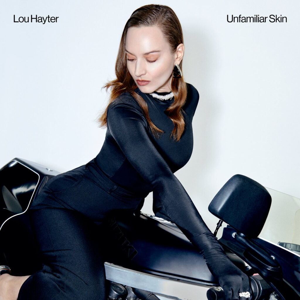 Lou Hayter in eleganter schwarzer Kleidung auf einem Motorrad – Cover zum 80er-inspirierten Electro-Pop-Album „Unfamiliar Skin“.