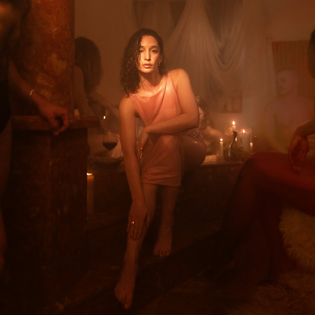 Elkka sitzt in einem warm ausgeleuchteten, sinnlichen Raum mit Kerzenlicht und anderen Körpern – das stimmungsvolle Cover von „Prism of Pleasure“.