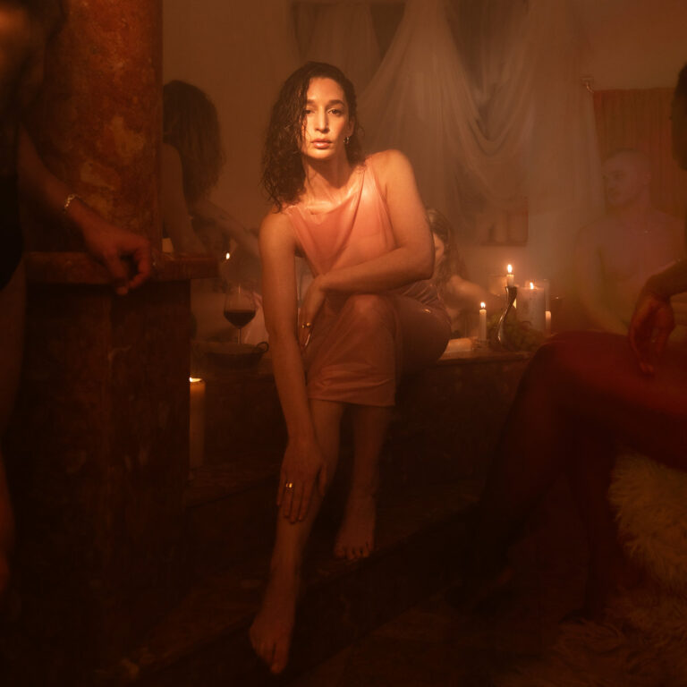 Elkka sitzt in einem warm ausgeleuchteten, sinnlichen Raum mit Kerzenlicht und anderen Körpern – das stimmungsvolle Cover von „Prism of Pleasure“.