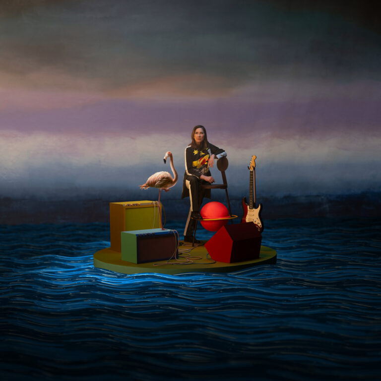 Kim Deal sitzt auf einer surrealen Plattform im Wasser, flankiert von E-Gitarre, Flamingo und skurrilen Objekten – Artwork des Albums „Nobody Loves You More“.