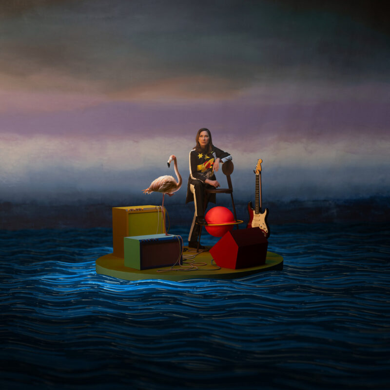 Kim Deal sitzt auf einer surrealen Plattform im Wasser, flankiert von E-Gitarre, Flamingo und skurrilen Objekten – Artwork des Albums „Nobody Loves You More“.