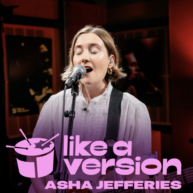 Asha Jefferies singt live ins Mikrofon bei einem Like a Version-Auftritt, mit geschlossenen Augen und weißer Rüschenbluse vor rotem Studiolicht.