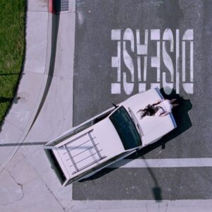 Lady Gaga liegt dramatisch auf einem weißen Wagen, der auf einer Straße mit dem Wort „DISEASE“ parkt – Szene aus dem Musikvideo zur Single.