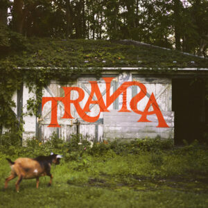 Ziege läuft über eine verwilderte Wiese vor einer alten Hütte mit großem roten Schriftzug „TRANSA“ – Albumcover mit ländlich-mystischer Atmosphäre.