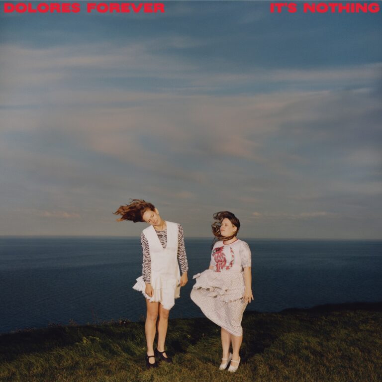 Cover von It’s Nothing von Dolores Forever: Zwei Frauen stehen im Wind auf einer Klippe mit Meer im Hintergrund, der Himmel weit und pastellfarben – stimmungsvoll und frei.