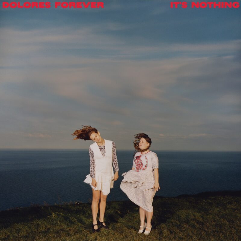 Cover von It’s Nothing von Dolores Forever: Zwei Frauen stehen im Wind auf einer Klippe mit Meer im Hintergrund, der Himmel weit und pastellfarben – stimmungsvoll und frei.