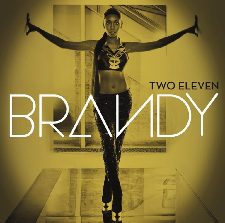 Brandy steht mit ausgestreckten Armen in einem modernen Raum, trägt ein bauchfreies Top und glänzende Hosen.