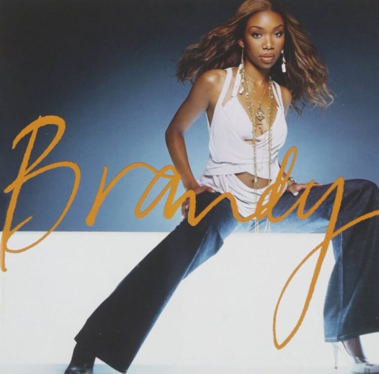 Brandy sitzt in lässiger Pose auf einem weißen Podest, trägt ein weißes Top und Jeans.
